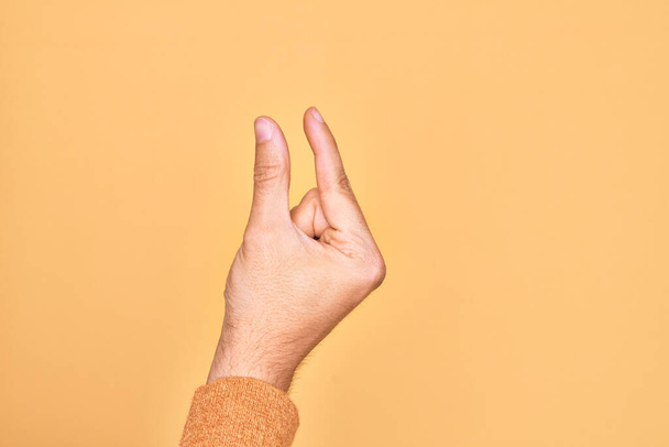 Hand eines kaukasischen jungen Mannes mit Fingern über isoliertem, gelbem Hintergrund, der unsichtbare Dinge pflückt und nimmt, Gegenstand mit Fingern hält, die Raum zeigen - Foto, Bild