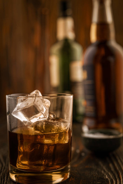 A glass of whiskey with ice, in the background are bottles on a wooden table of a bar counter, shallow depth of field, selective focus. The concept of alcoholic drinks in a roadside bar. - Photo, Image