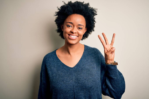 Junge schöne Afro-Amerikanerin mit lockigem Haar trägt lässigen Pullover und zeigt mit Finger Nummer drei nach oben, während sie selbstbewusst und glücklich lächelt. - Foto, Bild