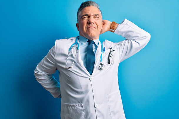 Middle age handsome grey-haired doctor man wearing coat and blue stethoscope Suffering of neck ache injury, touching neck with hand, muscular pain - Фото, изображение