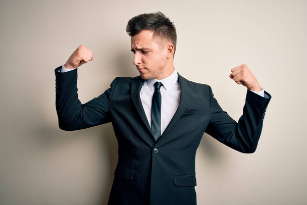 Молодой привлекательный бизнесмен в элегантном костюме и галстуке на изолированном фоне показывает мышцы рук, улыбающиеся с гордостью. Фитнес-концепция
. - Фото, изображение