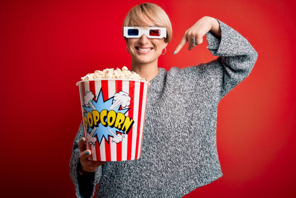 Fiatal szőke nő rövid haj visel 3D-s film szemüveg és eszik popcorn a moziban meglepett arccal mutogatva magára - Fotó, kép