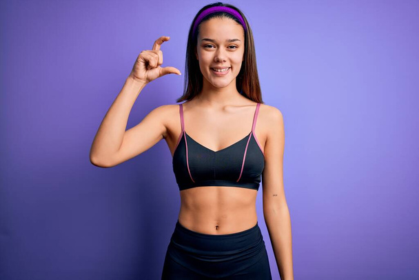 Young beautiful sporty girl doing sport wearing sportswear over isolated purple background smiling and confident gesturing with hand doing small size sign with fingers looking and the camera. Measure concept. - Photo, Image