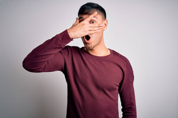 Jonge knappe Latijns-Amerikaanse man draagt casual shirt staan over witte geïsoleerde achtergrond gluren in shock bedekken gezicht en ogen met de hand, kijken door vingers met beschaamde uitdrukking. - Foto, afbeelding