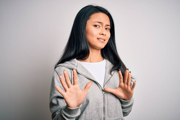 Junge schöne chinesische sportliche Frau trägt Sweatshirt über isoliertem weißem Hintergrund angeekelten Ausdruck, unzufrieden und ängstlich dabei Ekel Gesicht, weil Abneigung Reaktion. Mit erhobenen Händen - Foto, Bild