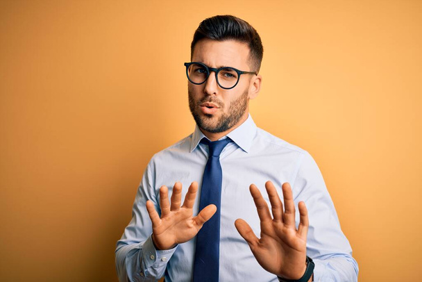 Young handsome businessman wearing tie and glasses standing over yellow background Moving away hands palms showing refusal and denial with afraid and disgusting expression. Stop and forbidden. - Photo, Image