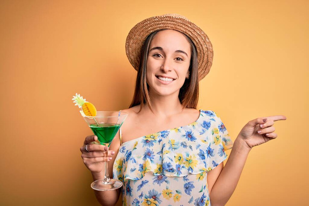 Νεαρή όμορφη τουρίστρια σε διακοπές φορώντας καλοκαιρινό καπέλο πίνοντας ποτό κοκτέιλ πολύ χαρούμενος δείχνοντας με το χέρι και το δάχτυλο στο πλάι - Φωτογραφία, εικόνα