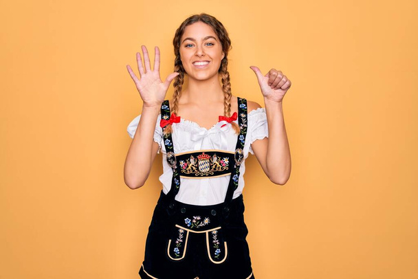 Junge schöne blonde deutsche Frau mit blauen Augen in traditionellem Oktoberfestkleid zeigt und zeigt mit Finger Nummer sechs nach oben, während sie selbstbewusst und glücklich lächelt. - Foto, Bild