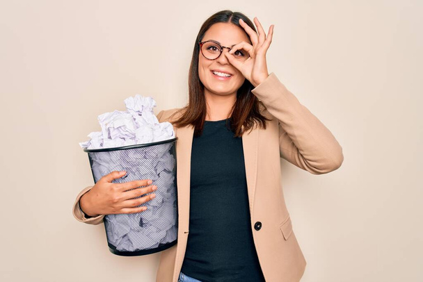 Молодая красивая брюнетка бизнес-женщина держит полный ящик бумаги калека бумаги улыбаясь счастливо делает хорошо знак с рукой на глазу глядя сквозь пальцы
 - Фото, изображение