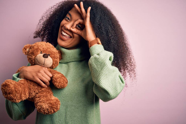 Young african american woman with afro hair hugging teddy bear over pink background with happy face smiling doing ok sign with hand on eye looking through fingers - 写真・画像