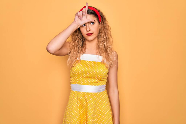 Beautiful blonde pin-up woman with blue eyes wearing diadem standing over yellow background making fun of people with fingers on forehead doing loser gesture mocking and insulting. - Foto, imagen