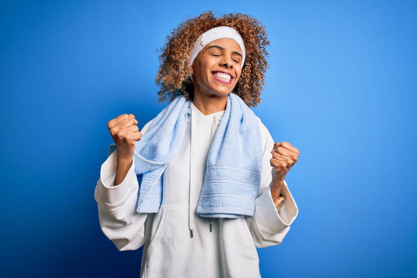 Jovem esportista afro-americana fazendo esporte vestindo roupas esportivas e toalha muito feliz e animado fazendo gesto vencedor com os braços levantados, sorrindo e gritando por sucesso. Conceito de celebração
. - Foto, Imagem