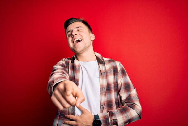 Giovane uomo caucasico bello indossa casual camicia moderna su sfondo rosso isolato ridere di te, indicando il dito verso la fotocamera con mano sul corpo, espressione di vergogna
 - Foto, immagini
