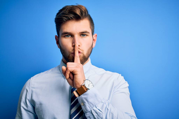 Young blond businessman with beard and blue eyes wearing elegant shirt and tie standing asking to be quiet with finger on lips. Silence and secret concept. - 写真・画像
