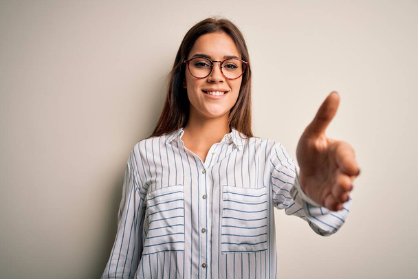 Young beautiful brunette woman wearing casual shirt and glasses over white background smiling friendly offering handshake as greeting and welcoming. Successful business. - Valokuva, kuva