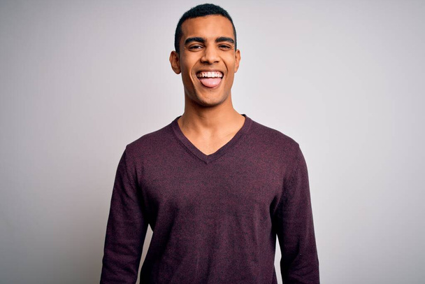 Jeune bel homme afro-américain portant un pull décontracté sur fond blanc collant la langue heureux avec une expression drôle. Concept d'émotion
. - Photo, image