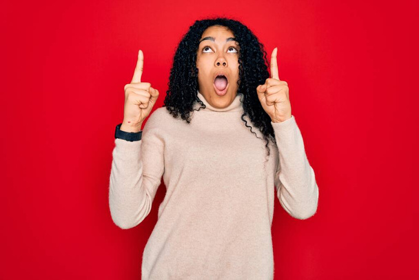 Mujer rizada afroamericana joven con suéter casual de cuello alto sobre fondo rojo asombrada y sorprendida mirando hacia arriba y señalando con los dedos y los brazos levantados
. - Foto, imagen