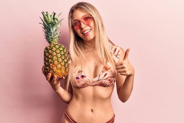 Mladá krásná blondýna žena v bikinách a slunečních brýlích drží ananas s úsměvem šťastný a pozitivní, palec nahoru dělá vynikající a schvalovací znamení  - Fotografie, Obrázek