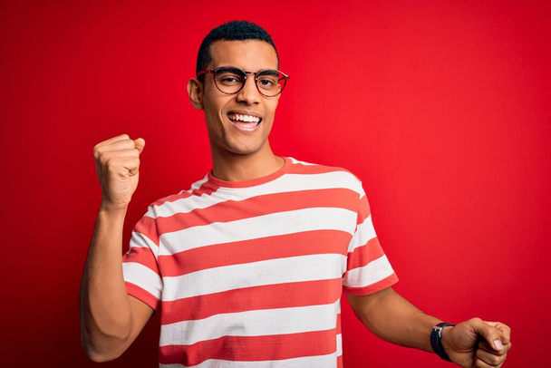 Junger gutaussehender afrikanisch-amerikanischer Mann in lässigem gestreiftem T-Shirt und Brille, fröhlich und fröhlich tanzend, lächelnd, lässig und selbstbewusst Musik hörend - Foto, Bild