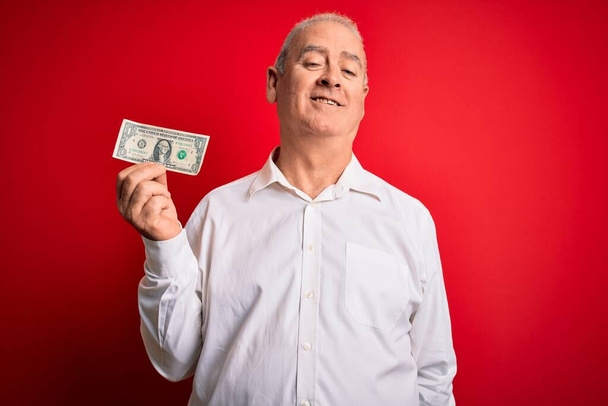 Middle age hoary man holding one dollar banknote over isolated red background with a happy face standing and smiling with a confident smile showing teeth - Valokuva, kuva