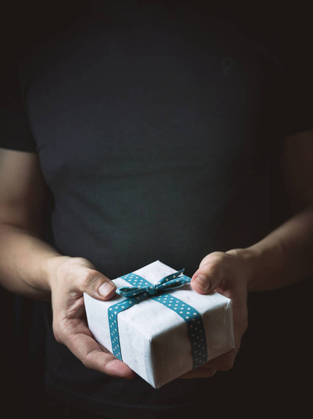 Primer plano de las manos masculinas sosteniendo un pequeño regalo envuelto con cinta azul. Un pequeño regalo en las manos de un hombre sobre fondo negro. Concéntrate en la pequeña caja. - Foto, Imagen