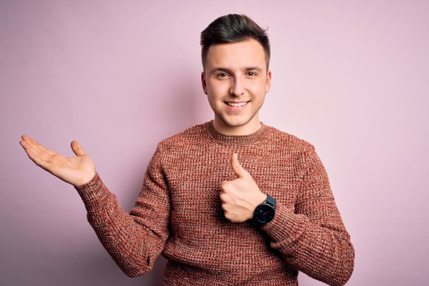 Jonge knappe blanke man draagt casual winter trui over roze geïsoleerde achtergrond Toont palm hand en doen ok gebaar met duimen omhoog, glimlachen gelukkig en vrolijk - Foto, afbeelding