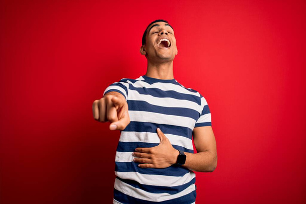 Bel homme afro-américain portant un t-shirt rayé décontracté debout sur fond rouge se moquant de vous, pointant du doigt l'appareil photo avec la main sur le corps, expression de honte
 - Photo, image