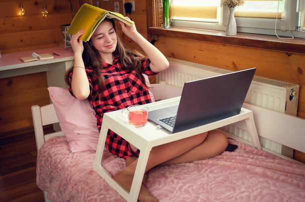Νεαρή μαθήτρια μαθαίνει μαθήματα σε απόσταση στο σπίτι σε ένα άνετο ξύλινο εσωτερικό στο κρεβάτι. - Φωτογραφία, εικόνα