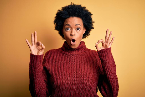 Young beautiful African American afro woman with curly hair wearing casual turtleneck sweater looking surprised and shocked doing ok approval symbol with fingers. Crazy expression - Zdjęcie, obraz