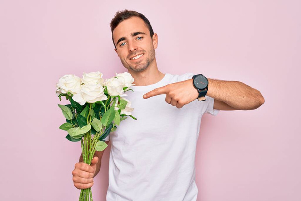 Mladý pohledný muž s modrýma očima drží kytice květin přes růžové pozadí velmi šťastný ukazující s rukou a prstem - Fotografie, Obrázek