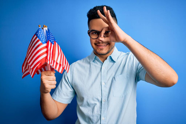 Jonge knappe patriottische man die de vlag van de Verenigde Staten vasthoudt en de onafhankelijkheidsdag viert met een vrolijk gezicht glimlachend, ok teken met hand op oog kijkend door de vingers - Foto, afbeelding