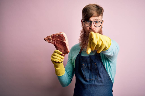 Ryšavý irský řezník s plnovousem držící syrový hovězí steak na růžovém pozadí ukazující prstem na fotoaparát a na vás, ruční znak, pozitivní a sebevědomé gesto zepředu - Fotografie, Obrázek