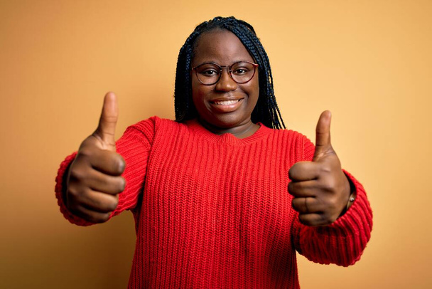 Mujer afroamericana de talla grande con trenzas que usan suéter casual sobre fondo amarillo aprobando hacer gesto positivo con la mano, pulgares arriba sonriendo y feliz por el éxito. Gesto del ganador
. - Foto, Imagen