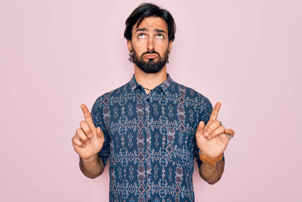 Jonge knappe Spaanse boheemse man draagt hippie stijl over roze achtergrond Wijzend omhoog kijken verdrietig en boos, richting met vingers, ongelukkig en depressief. - Foto, afbeelding