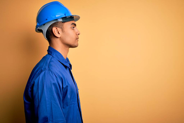 Νεαρός όμορφος Αφροαμερικάνος εργάτης φορώντας μπλε στολή και κράνος ασφαλείας κοιτάζοντας προς τα πλάγια, χαλαρώστε προφίλ ποζάρουν με φυσικό πρόσωπο με αυτοπεποίθηση χαμόγελο. - Φωτογραφία, εικόνα