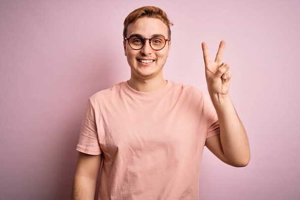 Fiatal jóképű, vörös hajú férfi alkalmi pólóban áll elszigetelt rózsaszín háttér felett, mutatva és mutatva a kettes számú ujjaival, miközben magabiztosan és boldogan mosolyog.. - Fotó, kép
