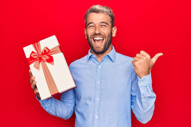 Junger schöner blonder Mann mit Bart hält Geburtstagsgeschenk über isoliertem rotem Hintergrund und zeigt mit dem Daumen zur Seite, lächelt glücklich mit offenem Mund - Foto, Bild