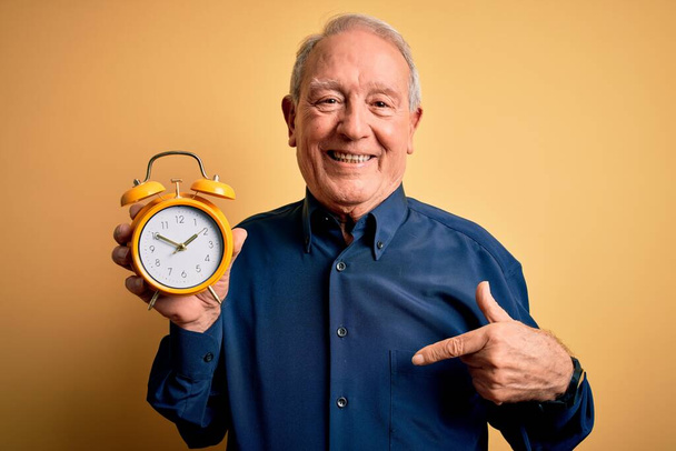 Senior grijze harige man met vintage wekker over gele achtergrond met verrassingsgezicht wijzend vinger naar zichzelf - Foto, afbeelding