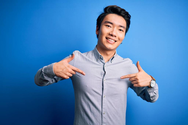 Młody przystojny Chińczyk ubrany w luźną koszulę stojący nad odizolowanym niebieskim tłem z uśmiechem na twarzy, wskazujący się palcami dumną i szczęśliwą. - Zdjęcie, obraz