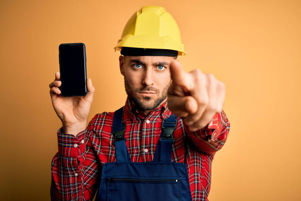 Молодий будівельник в шоломі безпеки, що показує екран смартфона на жовтому тлі, вказуючи пальцем на камеру і до вас, знак руки, позитивний і впевнений жест спереду
 - Фото, зображення