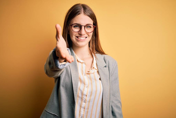 Young beautiful redhead woman wearing jacket and glasses over isolated yellow background smiling friendly offering handshake as greeting and welcoming. Successful business. - Valokuva, kuva