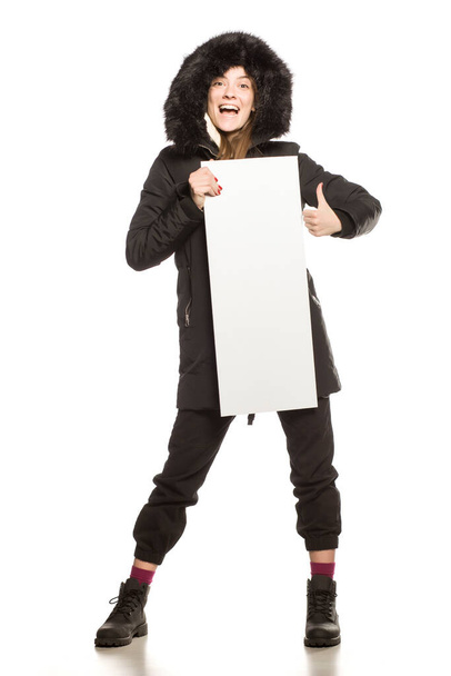Young model in winter jacket with hood shows an empty advertising board on white background - Foto, Imagen