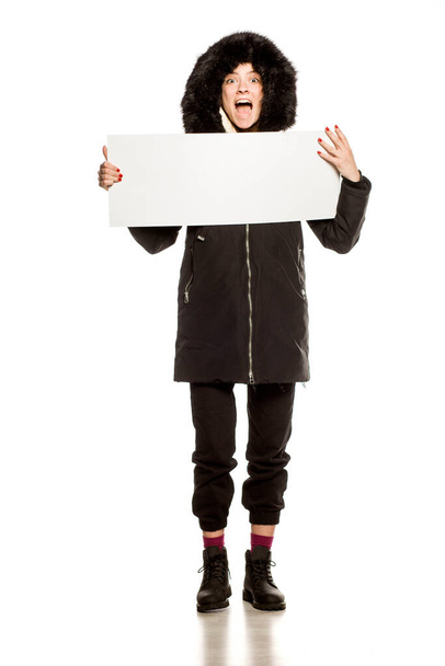 Jovem modelo feliz na jaqueta de inverno com capuz mostra uma placa de publicidade vazia no fundo branco
 - Foto, Imagem