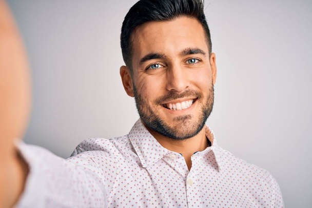 Νεαρός όμορφος άντρας με πουκάμισο που βγάζει σέλφι από την κάμερα σε λευκό φόντο με χαρούμενο πρόσωπο που στέκεται και χαμογελάει με αυτοπεποίθηση που δείχνει τα δόντια του - Φωτογραφία, εικόνα