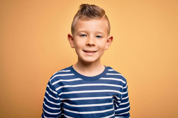 Young little caucasian kid with blue eyes wearing nautical striped shirt over yellow background with a happy and cool smile on face. Lucky person. - Fotoğraf, Görsel