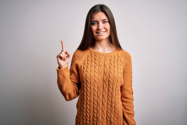 Young beautiful woman with blue eyes wearing casual sweater standing over white background with a big smile on face, pointing with hand finger to the side looking at the camera. - Foto, afbeelding