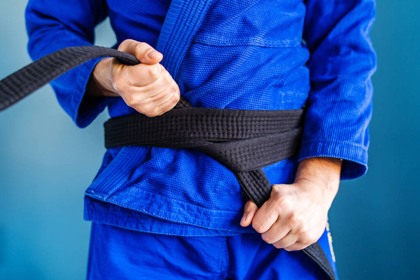 Close-up no meio da seção do jiu jitsu brasileiro bjj ou judo mãos cinto preto segurando e amarrar em torno da cintura de um lutador atleta vestindo kimono gi azul de pé na frente da parede
 - Foto, Imagem