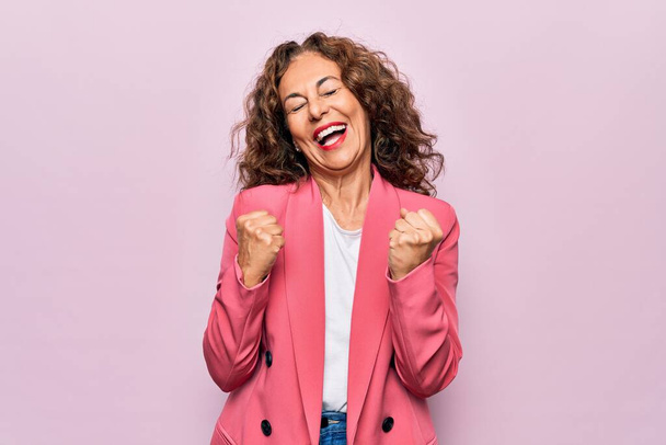 Mujer de mediana edad hermosa mujer de negocios con chaqueta de pie sobre fondo rosa aislado celebrando sorprendido y sorprendido por el éxito con los brazos levantados y los ojos cerrados
 - Foto, Imagen