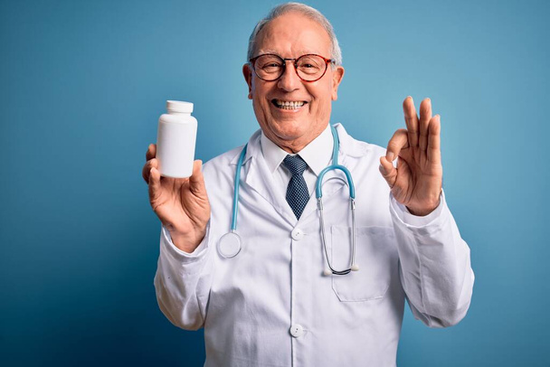 Senior gris doctor hombre sosteniendo píldoras farmacéuticas sobre fondo azul haciendo signo ok con los dedos, excelente símbolo
 - Foto, imagen