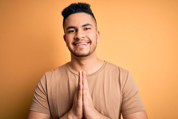 Young handsome man wearing casual t-shirt standing over isolated yellow background praying with hands together asking for forgiveness smiling confident. - Photo, Image